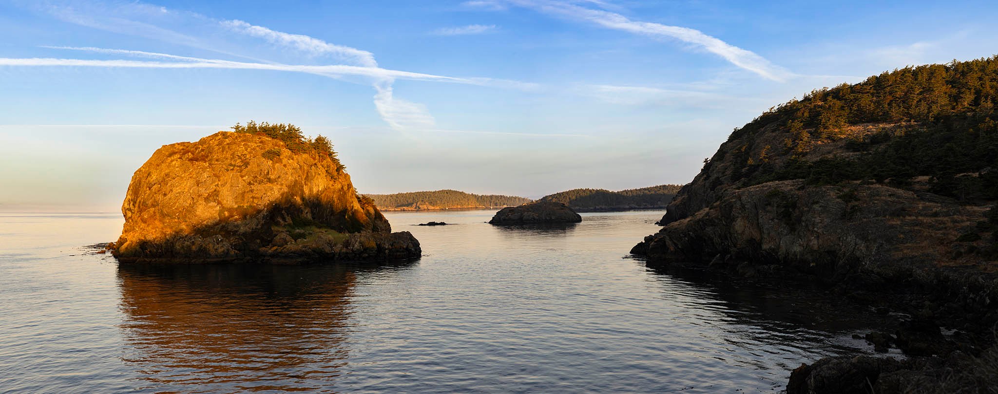 Rocky coastal cliffs and calm water at sunset in the San Juan Islands, with golden light illuminating the shoreline.