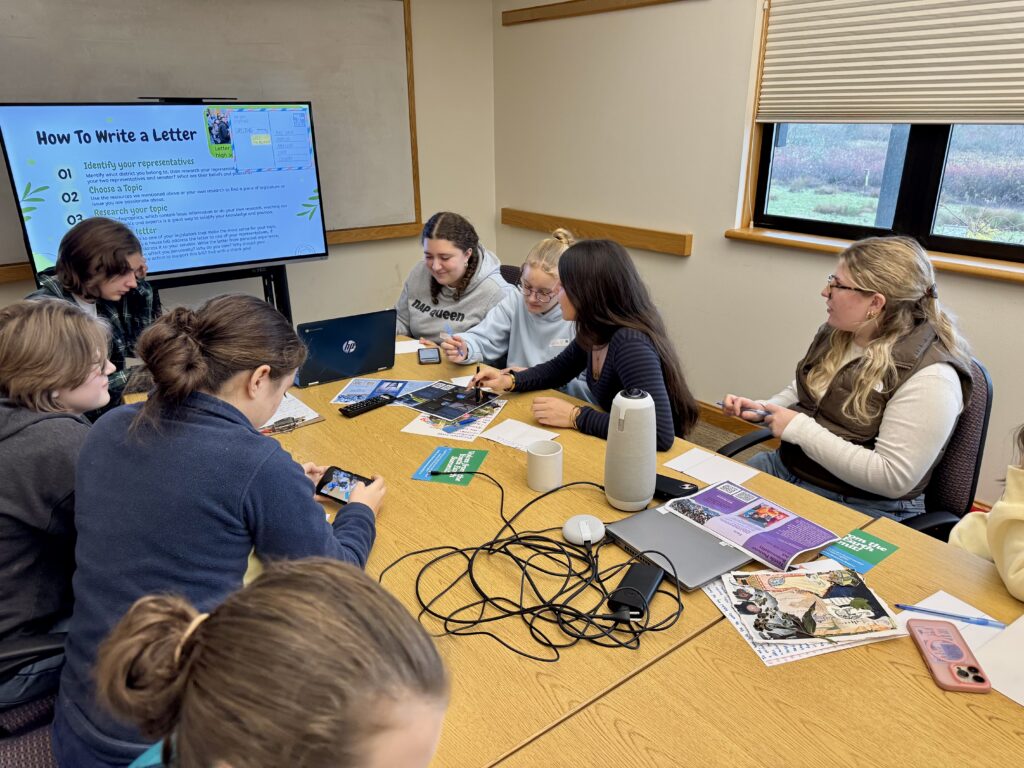 students sitting around a table, some writing, others looking at the screen with a powerpoint presentation titled "How to Write a Letter"